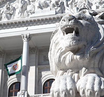 pormenor da fachada do Palácio de São Bento com estátua do leão no primeiro plano e, ao fundo, a varanda e a bandeira da Assembleia da República 